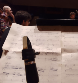 Image of a clarinet held up in front of some sheet music and an instrumental ensemble